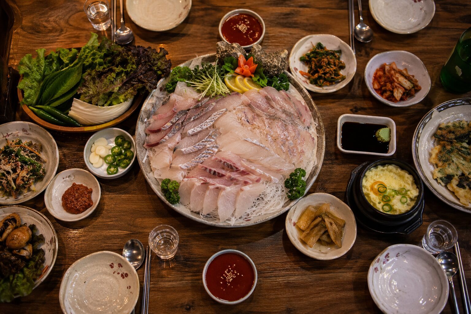 나무 테이블 가운데 회 한 접시, 둘레에 쌈 채소, 초장, 밑반찬, 소주잔